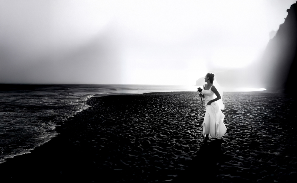 Bride in wedding dress stands alone on a rocky shore under a cloudy sky.