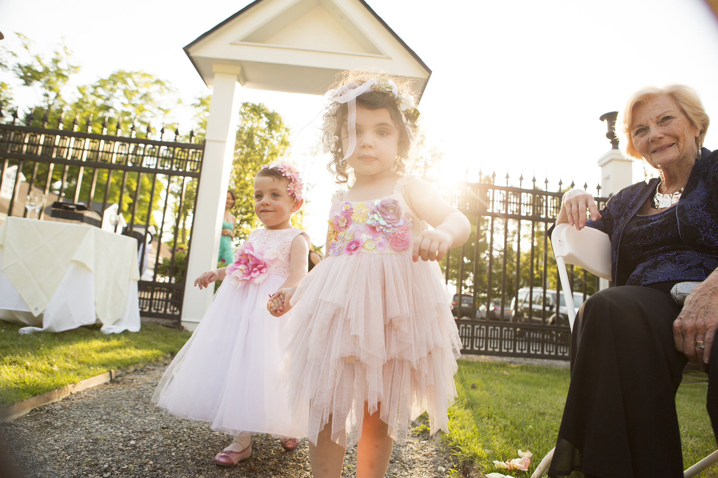 Two young girls in pastel dresses play outside near a white gazebo on a sunny day.