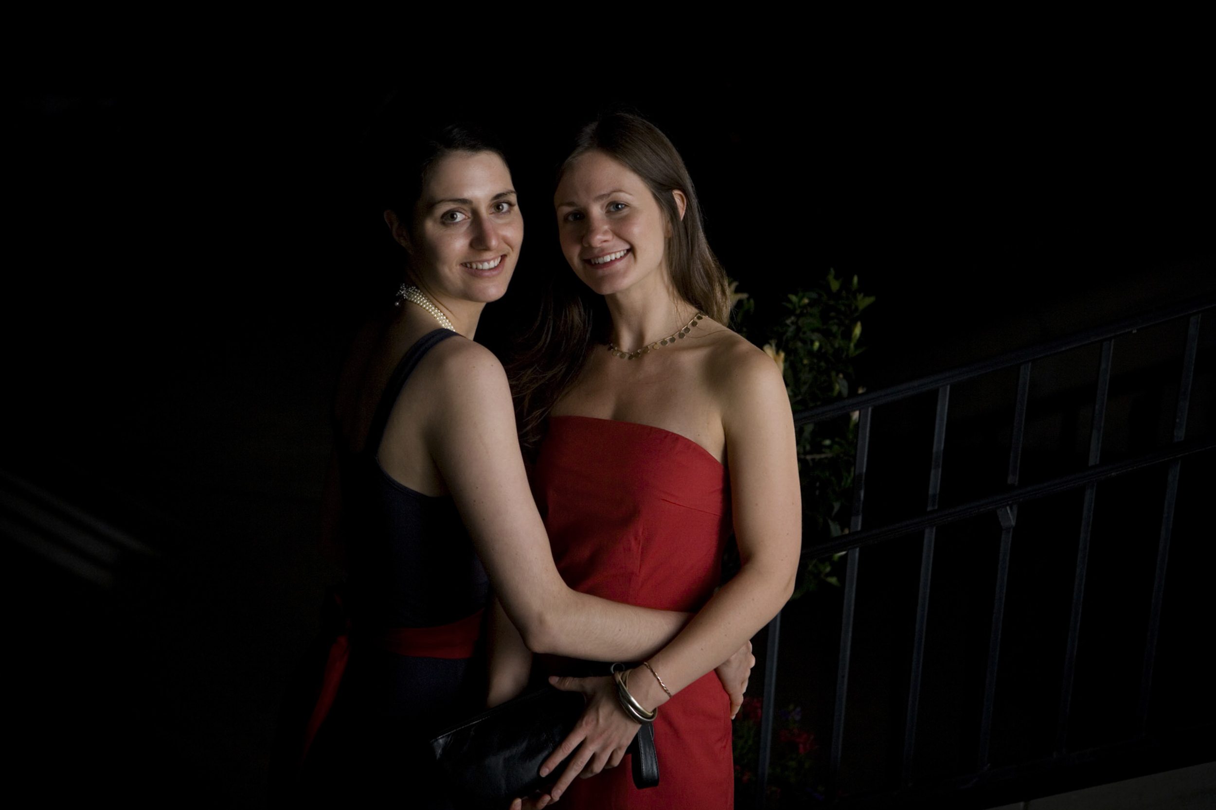 Two women dressed in elegant red dresses posing together in a dark setting.