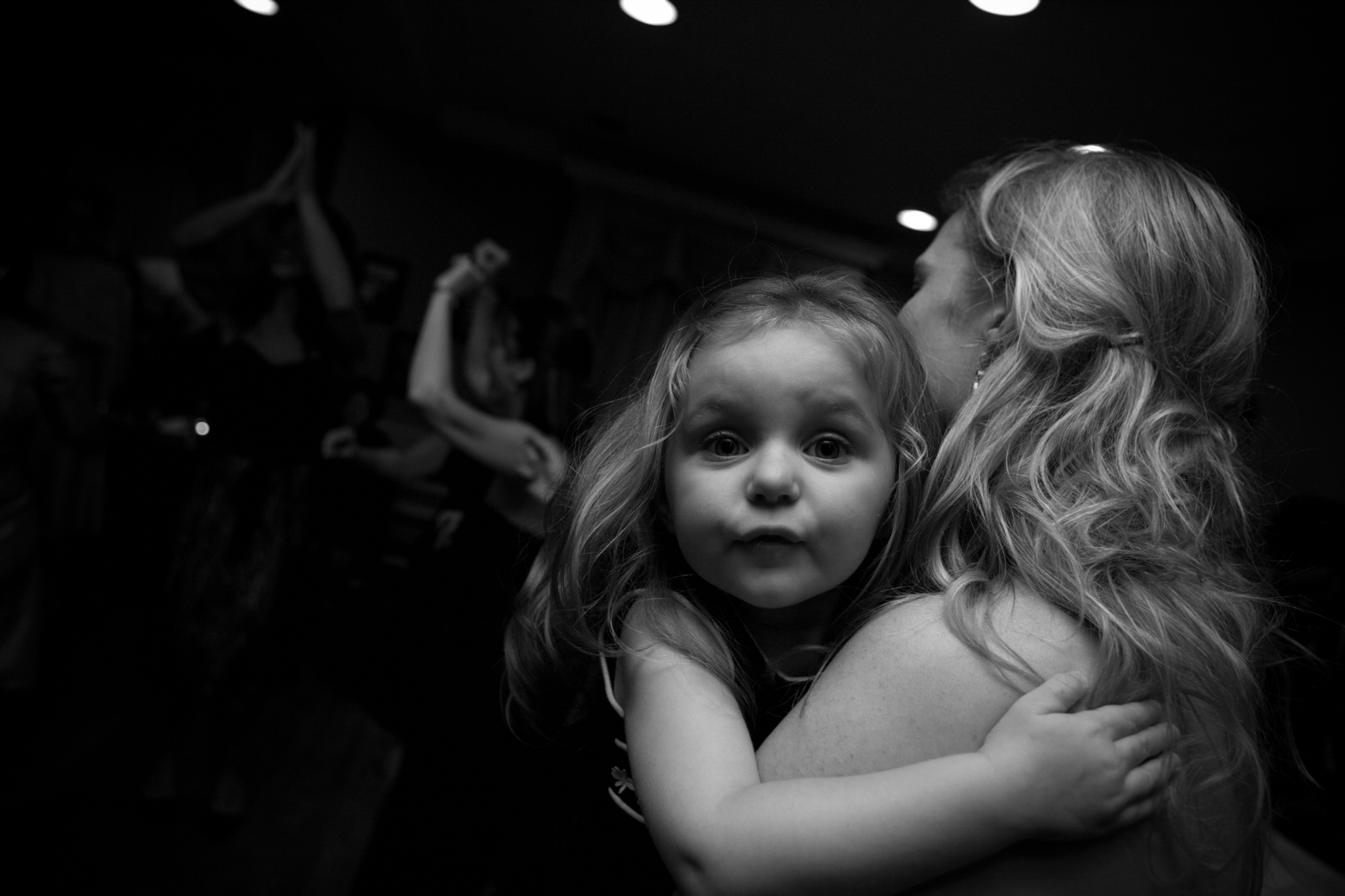 A young girl being held and looking curiously at the camera in a dimly lit room.