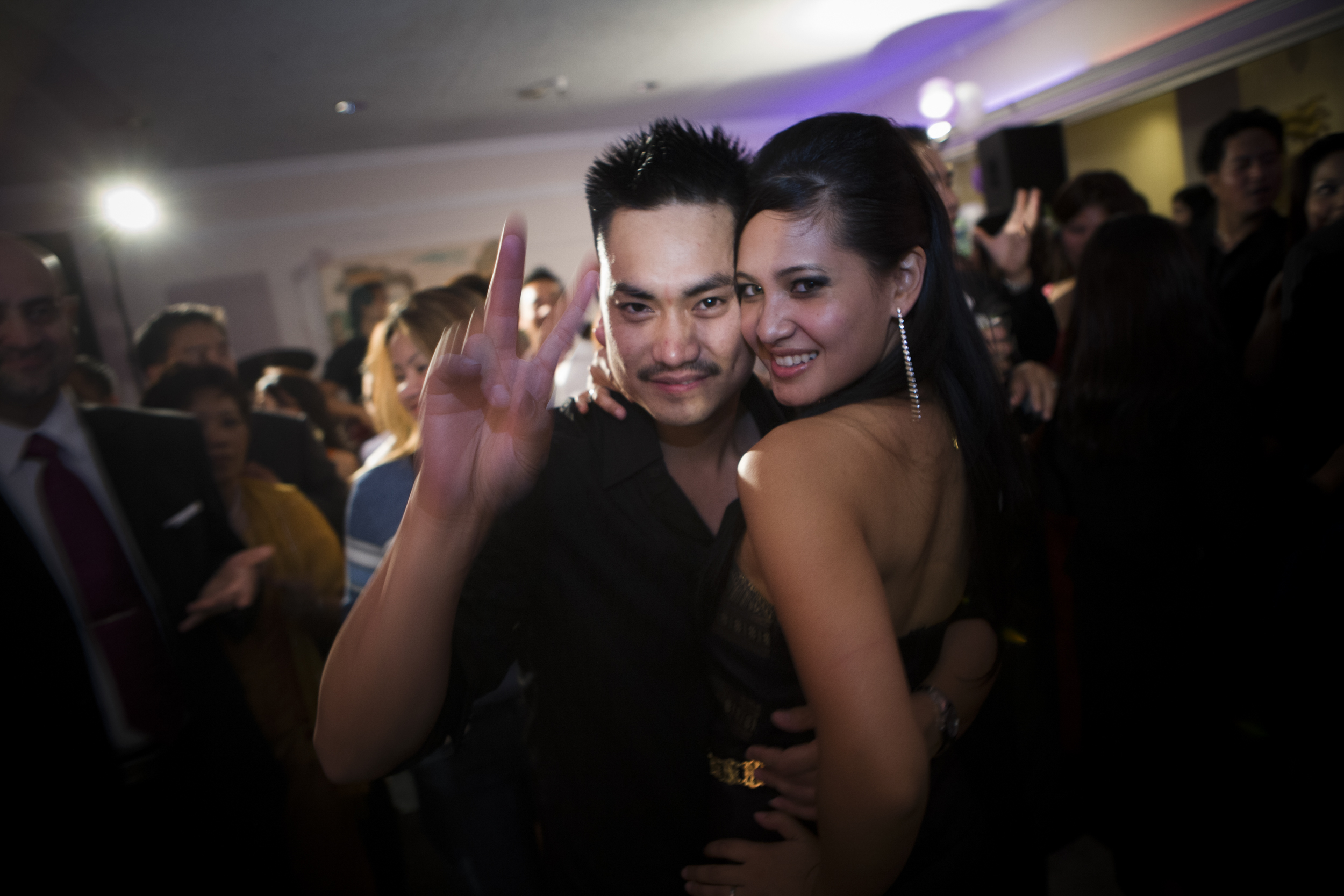 A happy couple posing at a lively party, with the man making a peace sign.