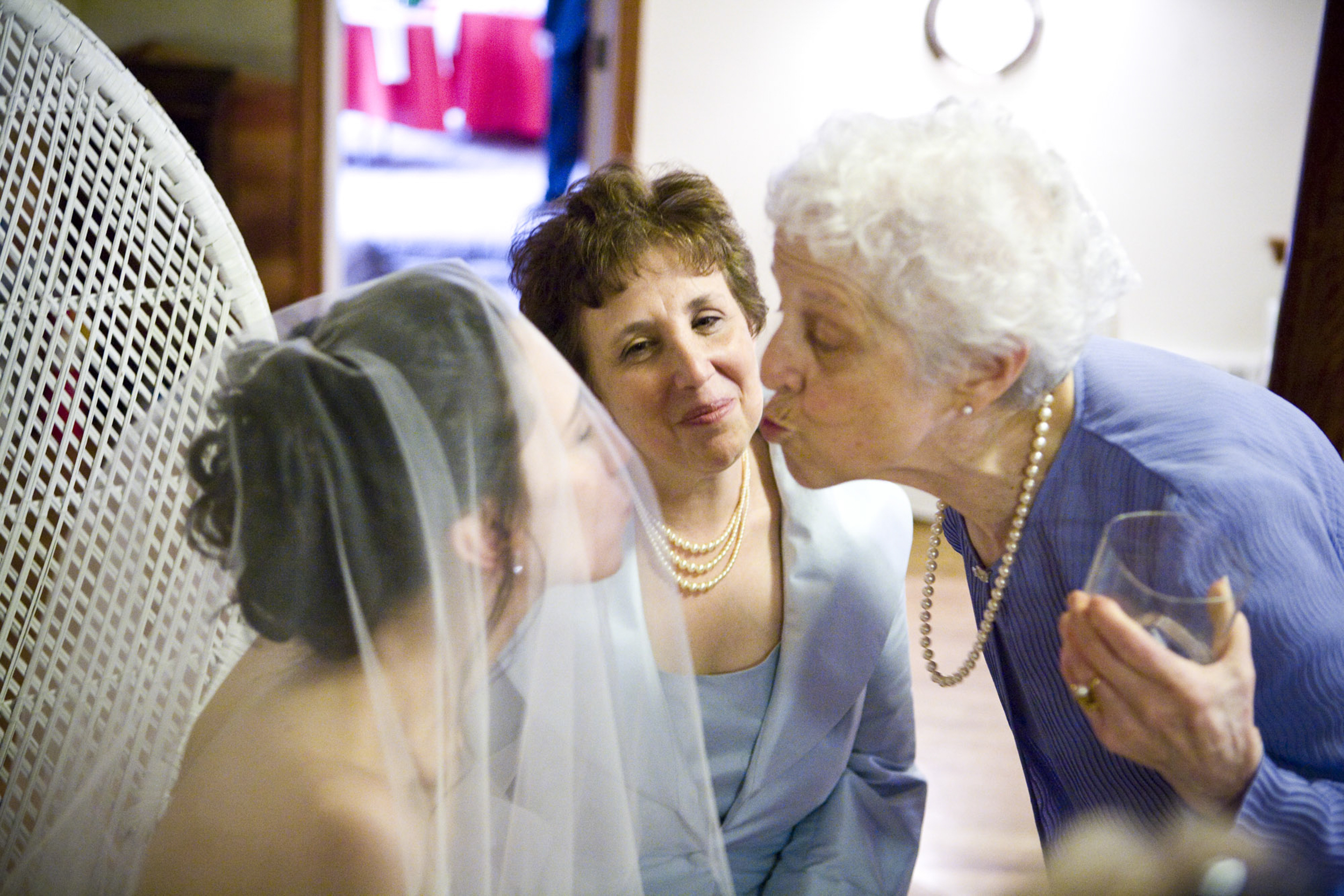 Elderly woman kisses bride while another woman watches warmly.