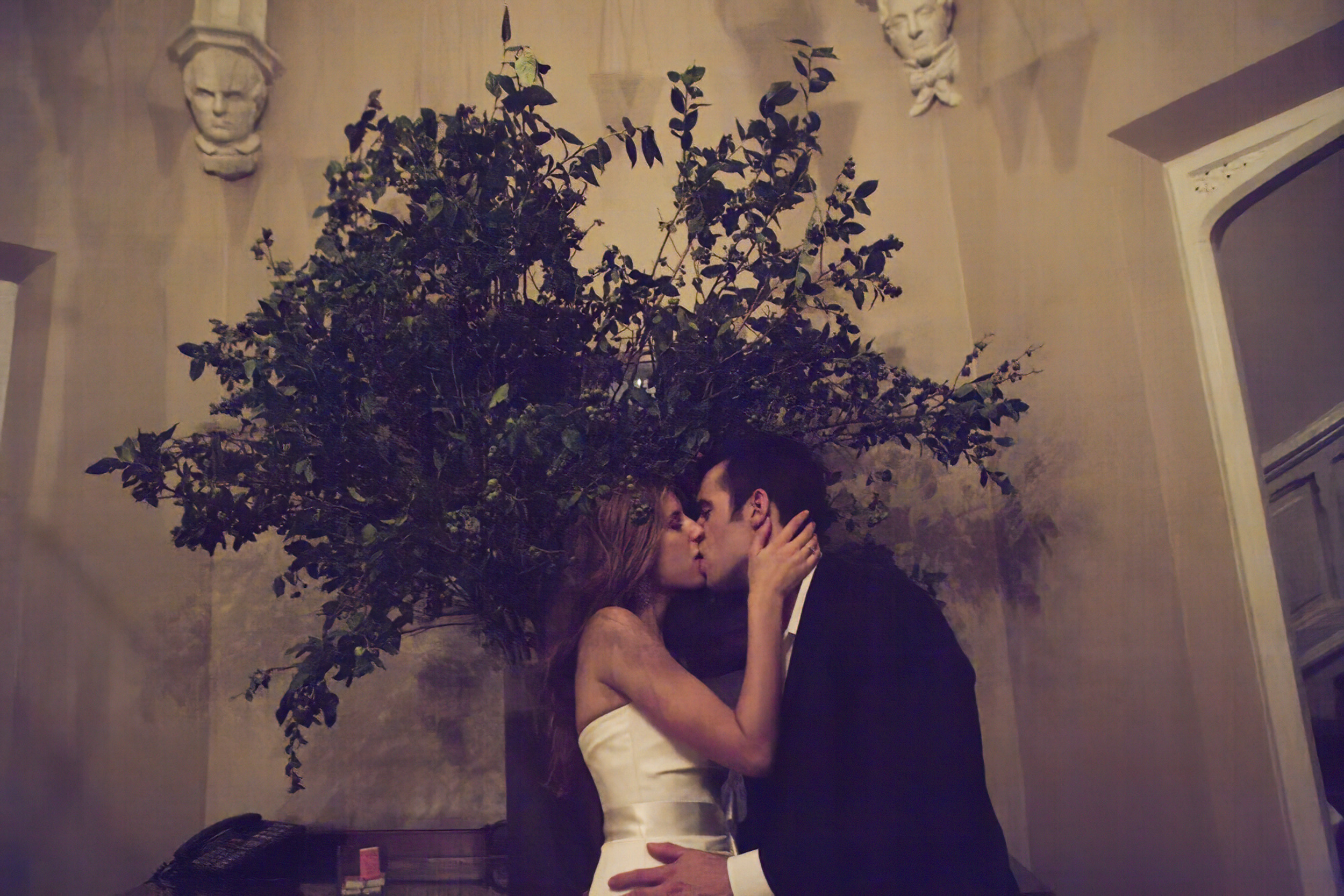 A couple shares a romantic kiss under dim lighting and greenery.