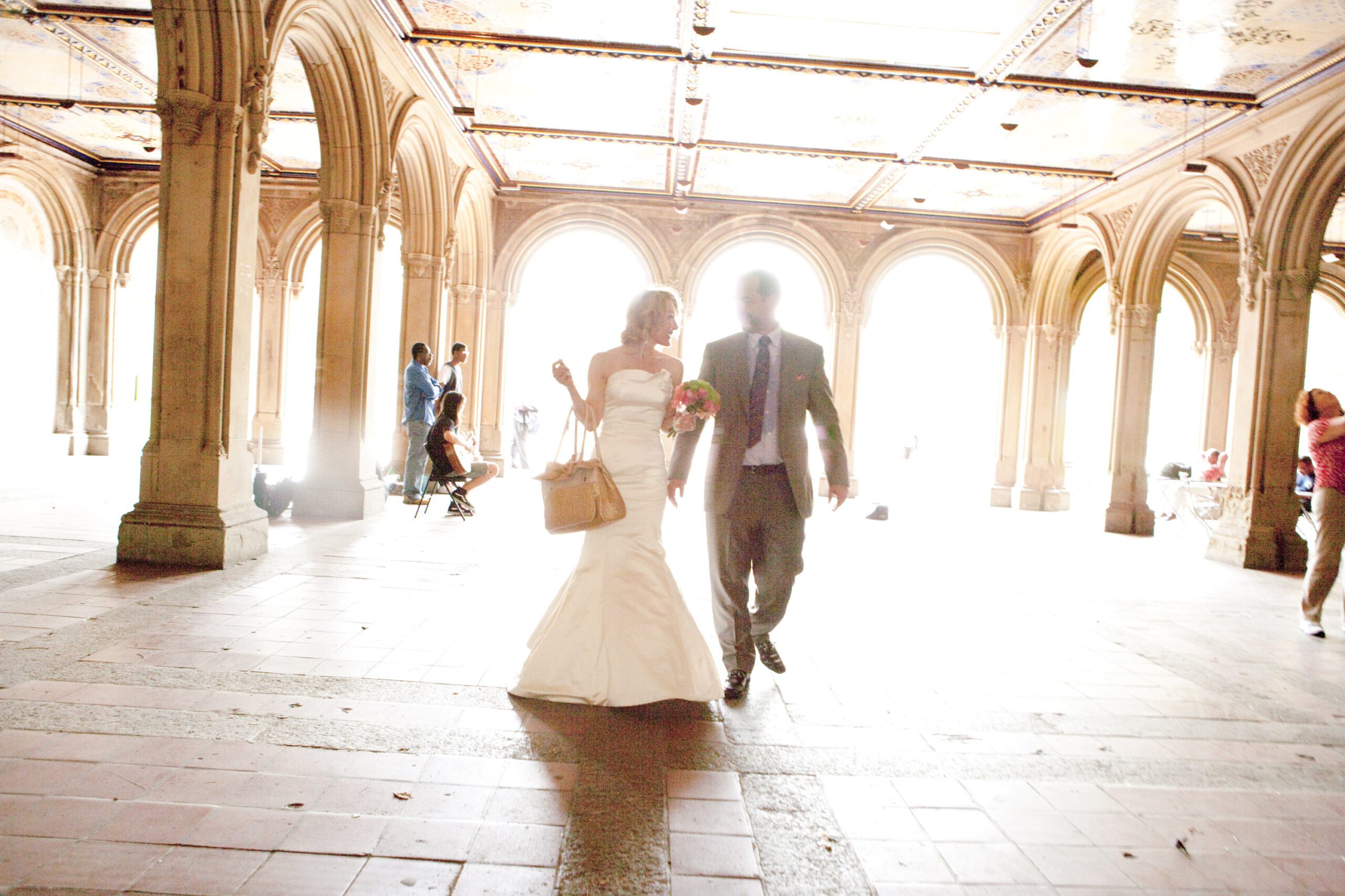 Bride and groom walking hand in hand in a sunlit archway.