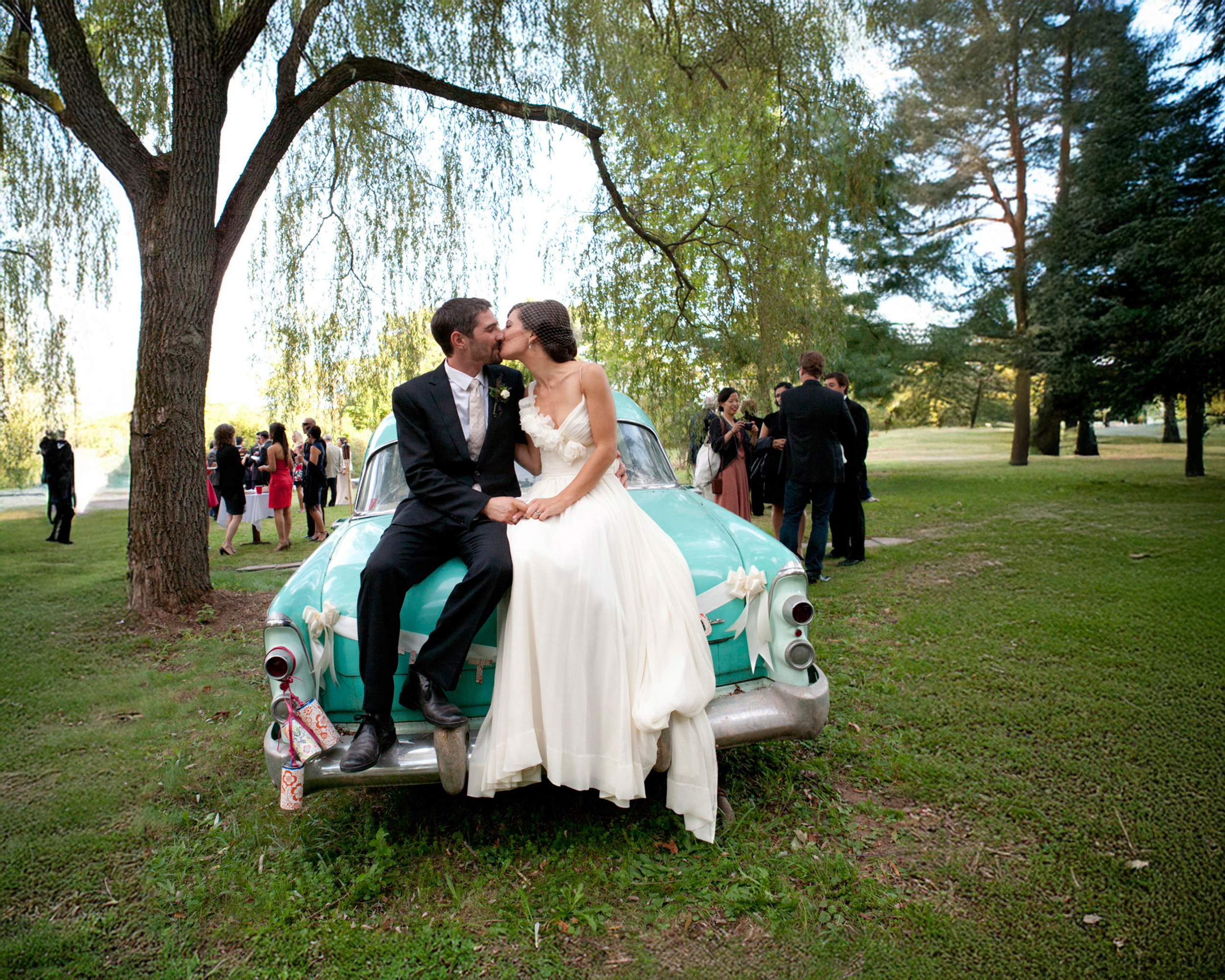 Newlyweds share a kiss while sitting on a vintage car at an outdoor wedding.