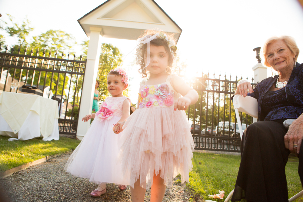 Two young girls dressed in floral and tulle dresses stand outdoors near a white gate.